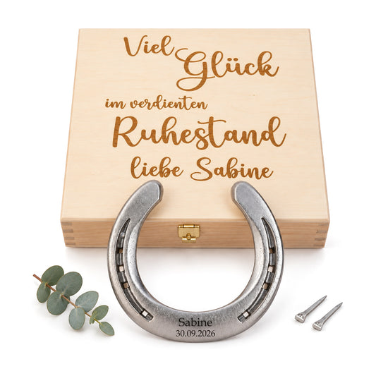 Glückshufeisen aus Stahl mit Gravur „Sabine 30.09.2026“ vor Holz-Geschenkbox mit Gravur „Viel Glück im verdienten Ruhestand“, Eukalyptuszweig und zwei Hufnägeln
